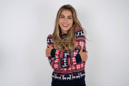Foto de Encantadora hermosa mujer rubia complacida que usa suéter de Navidad de punto abraza su propio cuerpo, se siente agradablemente cómodo posa. Concepto de ternura y autoestima - Imagen libre de derechos
