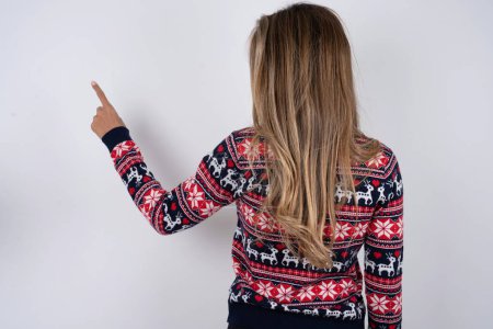 Téléchargez les photos : Belle femme blonde portant un pull de Noël tricoté pointant vers l'objet sur l'espace de copie, vue arrière. Tourne le dos - en image libre de droit