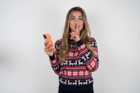 Foto de Hermosa mujer rubia vistiendo punto suéter de Navidad celebración de gadget moderno pedir no contar secretos - Imagen libre de derechos
