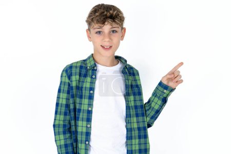 guapo adolescente chico usando camisa a cuadros sobre fondo blanco mirando a la cámara que indica dedo vacío espacio ventas