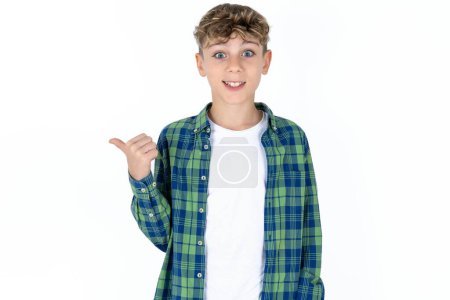 Téléchargez les photos : Charmant adolescent beau garçon portant une chemise à carreaux sur fond blanc pointant de côté avec index, montrant à l'espace de copie avoir des nouvelles sur les bonnes affaires - en image libre de droit
