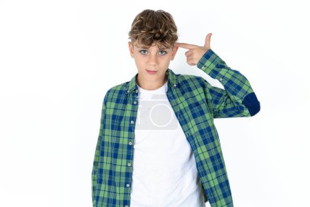Photo for Unhappy handsome teen boy wearing plaid shirt over white background makes suicide gesture and imitates gun with hand, curves lips keeps two fingers on temple, shoots, being tired of everything, - Royalty Free Image