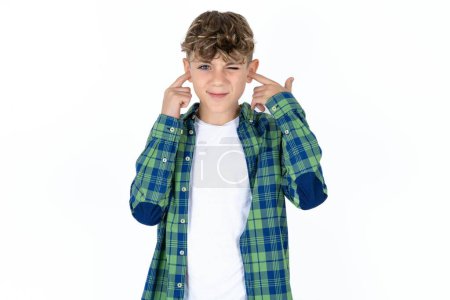 Téléchargez les photos : Beau adolescent garçon portant chemise à carreaux sur fond blanc couvrant les oreilles avec des doigts avec une expression agacée pour le bruit de la musique forte. Concept sourd. - en image libre de droit
