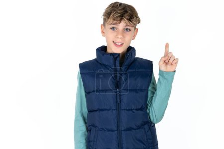 Foto de Guapo caucásico adolescente chico en azul chaleco mostrando y apuntando hacia arriba con el dedo número uno mientras sonríe confiado y feliz. - Imagen libre de derechos