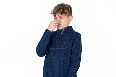 Téléchargez les photos : Beau garçon adolescent caucasien en pull bleu sentant quelque chose de puant et dégoûtant, odeur intolérable, retenant souffle avec les doigts sur le nez. Mauvaise odeur - en image libre de droit