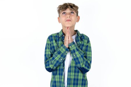 Téléchargez les photos : Beau adolescent garçon portant chemise à carreaux sur fond blanc mendier et prier avec les mains ainsi que l'expression de l'espoir sur le visage très émotionnel et inquiet. Mon Dieu ! - en image libre de droit