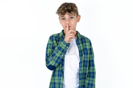 Foto de Guapo adolescente con camisa a cuadros sobre fondo blanco hace gesto de silencio, mantiene el dedo sobre los labios. Silencio y concepto secreto. - Imagen libre de derechos