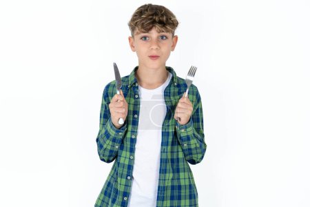 Foto de Hambriento guapo adolescente chico usando camisa a cuadros sobre fondo blanco sosteniendo en mano tenedor cuchillo quiere sabroso delicioso pastel de pizza - Imagen libre de derechos
