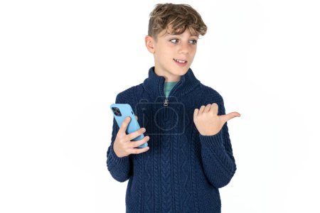 Téléchargez les photos : Beau caucasien adolescent garçon en bleu pull en utilisant et textos avec smartphone pointant et montrant avec pouce sur le côté avec heureux visage souriant - en image libre de droit