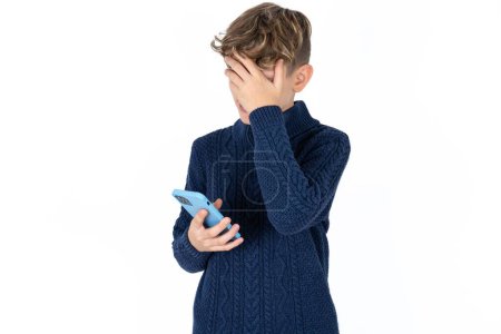 Photo for Handsome Caucasian teen boy in blue sweater looking at smartphone feeling sad holding hand on face. - Royalty Free Image