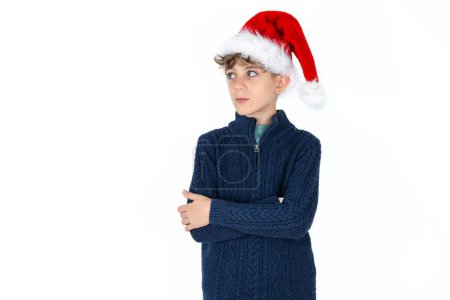 Téléchargez les photos : Charmant garçon adolescent caucasien élégant et réfléchi en pull bleu et chapeau de Noël se tient avec les bras repliés concentrés quelque part avec expression coûteuse pense quoi faire - en image libre de droit