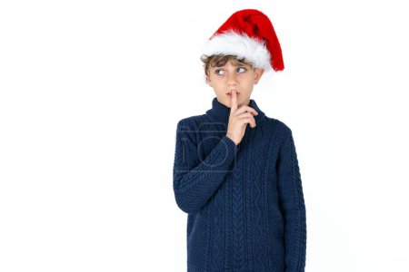 Téléchargez les photos : Beau garçon adolescent caucasien en pull bleu et chapeau de Noël geste de silence garde index aux lèvres fait signe de silence. Demande de ne pas partager le secret - en image libre de droit