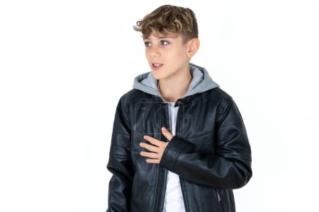 Photo for Joyful handsome teen boy in leather jacket posing over white studio background expresses positive emotions recalls something funny keeps hand on chest and giggles happily. - Royalty Free Image