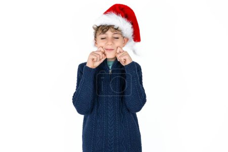 Photo for Pleased handsome caucasian teen boy in blue sweater and christmas hat with closed eyes keeps hands near cheeks and smiles tenderly imagines something very pleasant - Royalty Free Image