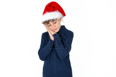 Foto de Guapo caucásico adolescente chico en azul suéter y navidad sombrero se apoya en prensado palmas cierra los ojos y tiene agradable sonrisa sueña con algo - Imagen libre de derechos