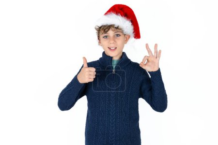 Foto de Guapo caucásico adolescente chico en azul suéter y sombrero de navidad sonriendo y buscando feliz, despreocupado y positivo, la victoria gesto o la paz con una mano - Imagen libre de derechos