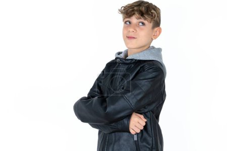 Foto de Guapo adolescente chico en cuero chaqueta posando sobre blanco estudio de fondo brazos cruzados mirada vacío espacio cartelera - Imagen libre de derechos