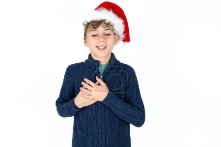 Foto de Guapo caucásico adolescente chico en azul suéter y navidad sombrero expresa felicidad, ríe agradablemente, mantiene las manos en el corazón - Imagen libre de derechos