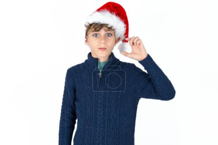 Téléchargez les photos : Beau garçon adolescent caucasien en pull bleu et chapeau de Noël porte-monnaie lèvre et gestes à la main, montre quelque chose de très peu. - en image libre de droit
