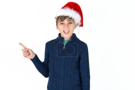 Téléchargez les photos : Beau garçon adolescent caucasien en pull bleu et chapeau de Noël pointe de côté sur copie espace vide. Promotion des personnes et concept de publicité - en image libre de droit