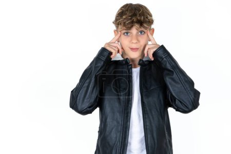 Photo for Serious concentrated handsome teen boy in leather jacket posing over white studio background keeps fingers on temples, tries to ease tension, gather with thoughts and remember important information for exam - Royalty Free Image