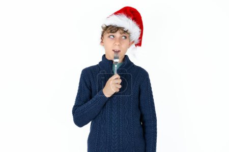 Foto de Foto de niño adolescente caucásico guapo de ensueño en suéter azul y sombrero de Navidad lamer tenedor mirar espacio vacío - Imagen libre de derechos