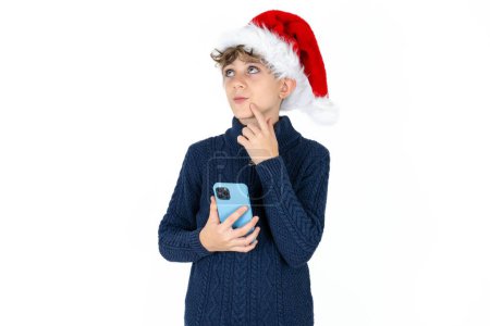 Photo for Handsome caucasian teen boy in blue sweater and christmas hat thinks deeply about something, uses modern mobile phone, tries to made up good message, keeps index finger near lips. - Royalty Free Image