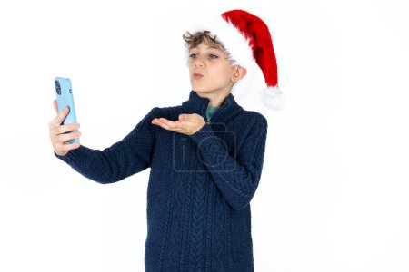 Téléchargez les photos : Beau garçon adolescent caucasien en pull bleu et chapeau de Noël souffle baiser d'air à la caméra du smartphone et prend selfie, envoie mwah via appel en ligne. - en image libre de droit