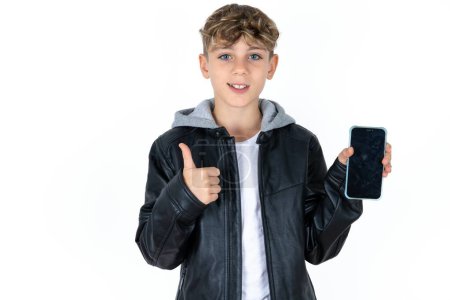 Foto de Guapo adolescente chico en chaqueta de cuero posando sobre fondo blanco estudio Mostrar pantalla en blanco smartphone, pulgar hacia arriba recomendar nueva aplicación - Imagen libre de derechos
