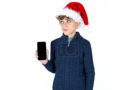 Téléchargez les photos : Beau garçon adolescent caucasien en pull bleu et chapeau de Noël tient un nouveau téléphone mobile et semble mystérieux de côté montre l'affichage vierge de cellulaire moderne - en image libre de droit