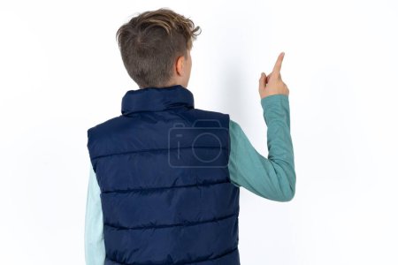 Téléchargez les photos : Beau garçon adolescent caucasien en gilet bleu pointant vers l'objet sur l'espace de copie, vue arrière. Tourne le dos - en image libre de droit