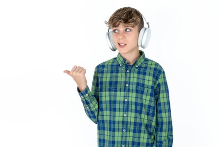 Photo for Handsome teen boy wearing plaid shirt over white background listens audio track via wireless headphones points thumb away advertises copy space - Royalty Free Image
