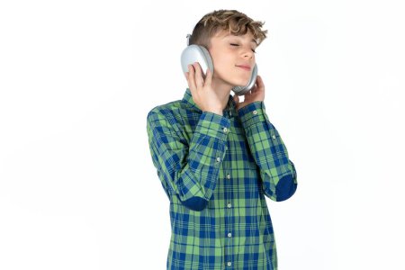 Photo for Handsome teen boy wearing plaid shirt over white background with headphones on her head, listens to music, enjoying favourite song with closed eyes, holding hands on headset. - Royalty Free Image