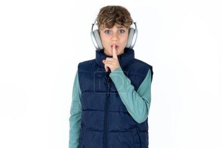 Photo for Handsome Caucasian teen boy in blue vest making hush gesture with finger on her lips wearing  wireless headphones. Be quiet. - Royalty Free Image