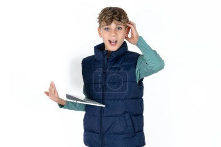 Foto de Foto de chico adolescente caucásico guapo sorprendido en chaleco azul que sostiene la cabeza del brazo del gadget moderno - Imagen libre de derechos