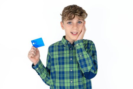 Foto de Asombroso positivo guapo adolescente con camisa a cuadros sobre fondo blanco con brazo cara sostener banco tarjeta de plástico funciones impresas - Imagen libre de derechos