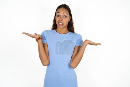 Téléchargez les photos : Clueless jeune belle femme hispanique portant un t-shirt bleu sur fond blanc hausse les épaules avec hésitation, fait face à une situation douteuse, écarte les paumes, Décision difficile - en image libre de droit