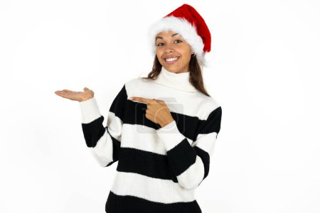 Photo for Young beautiful woman wearing striped sweater and a santa claus hat pointing and holding hand showing adverts - Royalty Free Image