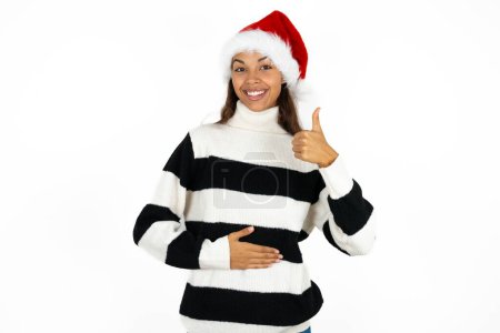 Foto de Joven hermosa mujer con suéter a rayas y un sombrero de Santa Claus feliz sonrisa positiva manos en el vientre muestran el pulgar hacia arriba bien saludable - Imagen libre de derechos