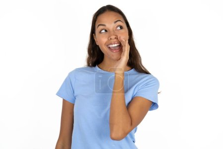 Foto de Joven hermosa mujer hispana vistiendo azul camiseta sobre fondo blanco escuchar noticias privadas increíbles impresionado grito compartir - Imagen libre de derechos
