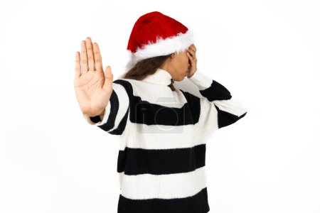 Photo for Young beautiful woman wearing striped sweater and a santa claus hat covers eyes with palm and doing stop gesture, tries to hide from everybody. - Royalty Free Image