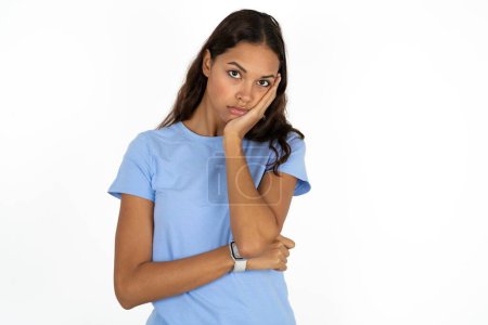 Foto de Joven mujer hispana hermosa muy aburrida usando una camiseta azul sobre fondo blanco agarrada de la mano en la mejilla mientras la apoya con otra mano cruzada, luciendo cansada y enferma, - Imagen libre de derechos