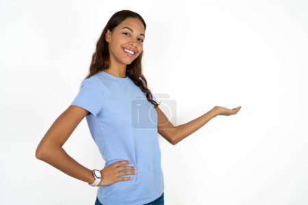 Foto de Joven hermosa mujer hispana vistiendo camiseta azul sobre fondo blanco sintiéndose feliz y alegre, sonriendo y dándote la bienvenida, invitándote con un gesto amistoso - Imagen libre de derechos