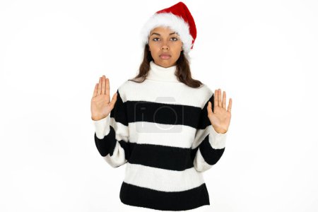 Photo for Serious Young beautiful woman wearing striped sweater and a santa claus hat pulls palms towards camera, makes stop gesture, asks to control your emotions and not be nervous - Royalty Free Image