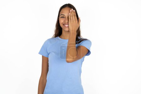 Téléchargez les photos : Jeune belle hispanique femme portant bleu t-shirt sur fond blanc couvrant un oeil avec sa main, sourire confiant sur le visage et l'émotion surprise. - en image libre de droit
