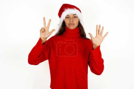 Foto de Joven hermosa mujer vistiendo suéter rojo y sombrero de Santa Claus en la Navidad mostrando y señalando hacia arriba con los dedos número ocho mientras sonríe confiado y feliz. - Imagen libre de derechos