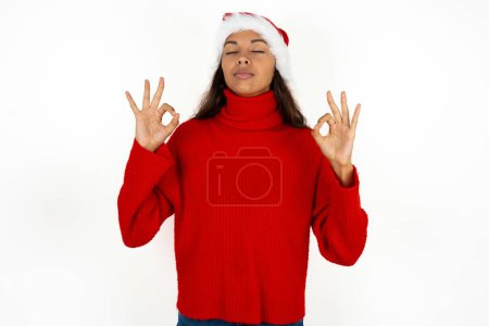 Photo for Young beautiful woman wearing red sweater and santa claus hat at christmas relax and smiling with eyes closed doing meditation gesture with fingers. Yoga concept. - Royalty Free Image