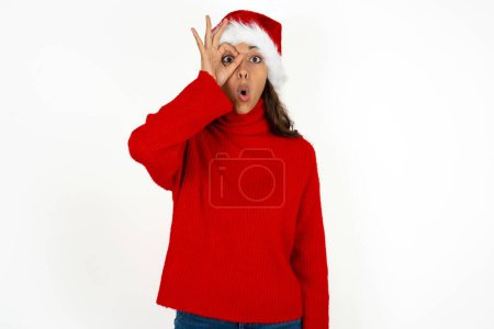 Téléchargez les photos : Jeune belle femme portant un pull rouge et santa claus chapeau à Noël faisant geste ok choqué par le visage surpris, oeil regardant à travers les doigts. Expression incrédule. - en image libre de droit