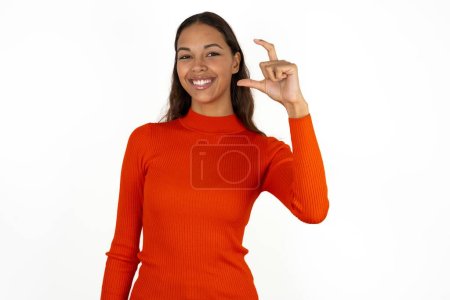 Téléchargez les photos : Jeune femme hispanique portant col roulé rouge sur fond blanc geste souriant et confiant avec la main faisant signe de petite taille avec les doigts regardant et la caméra. Concept de mesure - en image libre de droit