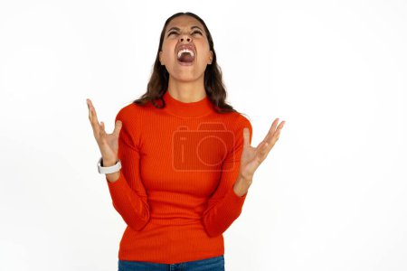 Foto de Mujer hispana joven con cuello alto rojo sobre fondo blanco loco y loco gritando y gritando con expresión agresiva y los brazos levantados. Concepto de frustración. - Imagen libre de derechos